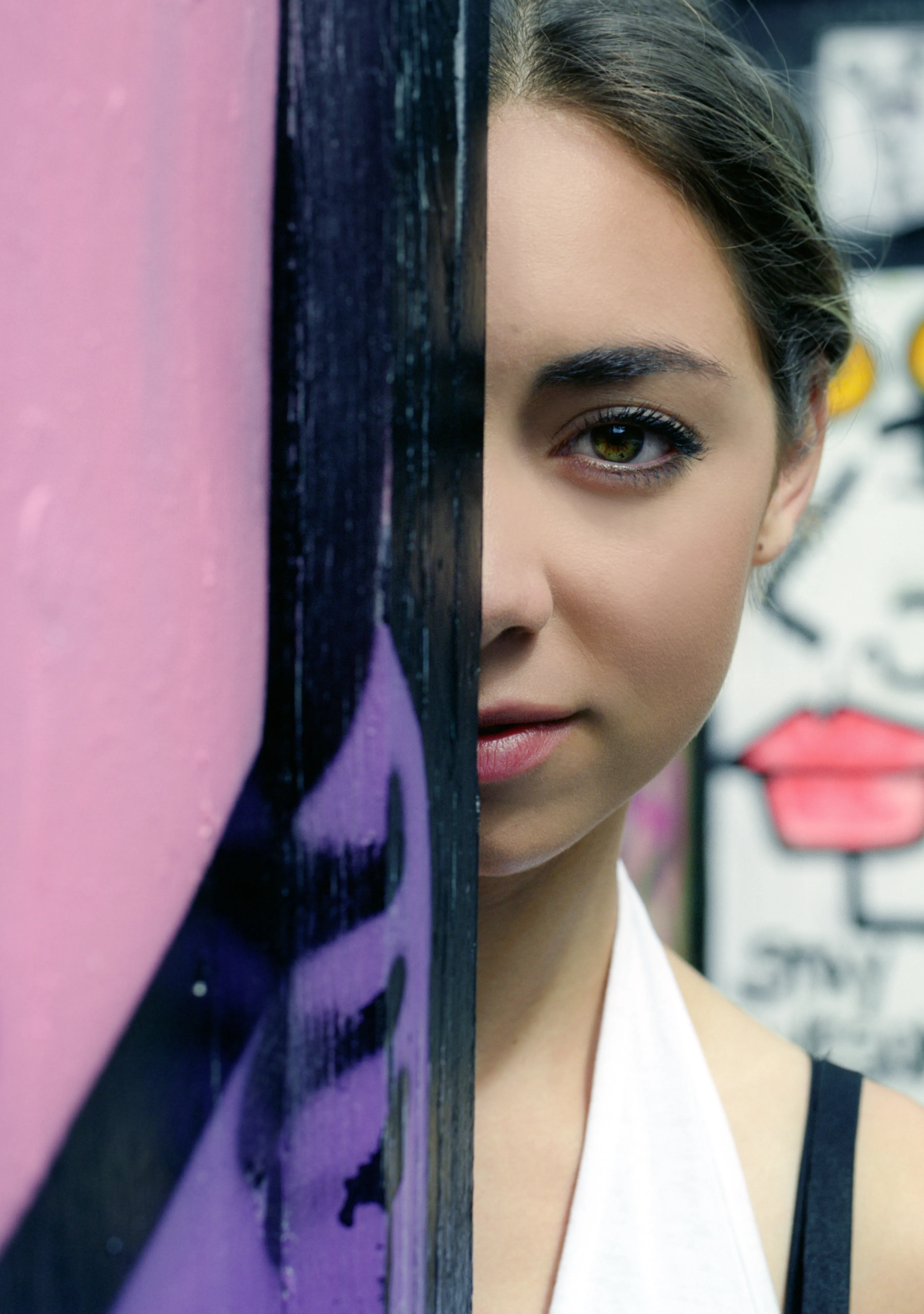Half of Riya's face is hidden behind the dark edge of a wall; her visible eye meets the camera, with a wall painted pink and purple to one side.