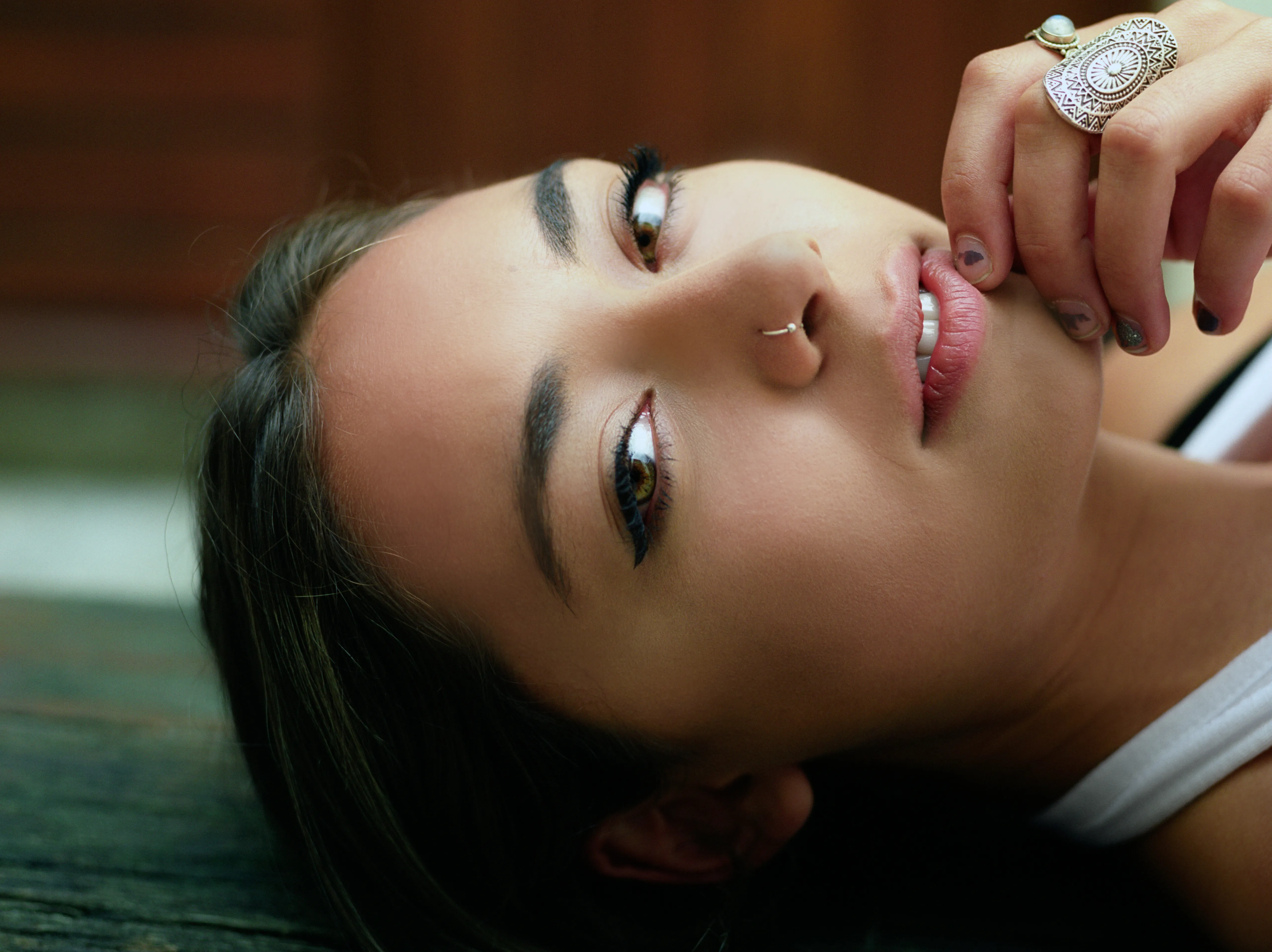 Riya lies on her side on a weathered wooden surface, one ringed hand raised near her mouth, looking up towards the camera.