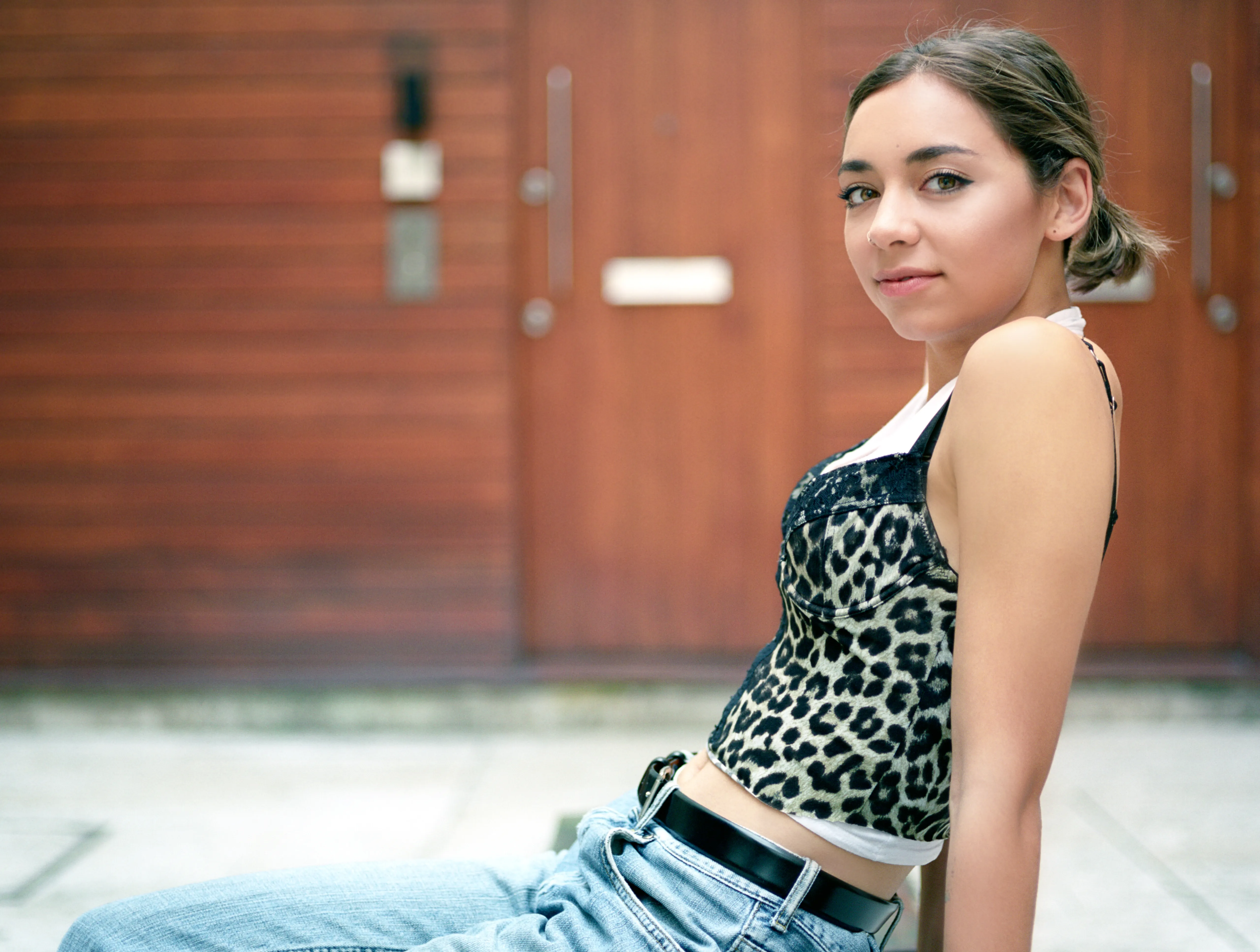 Riya sits on the ground leaning back against a wooden door, turning over her shoulder towards the camera with a slight smile.