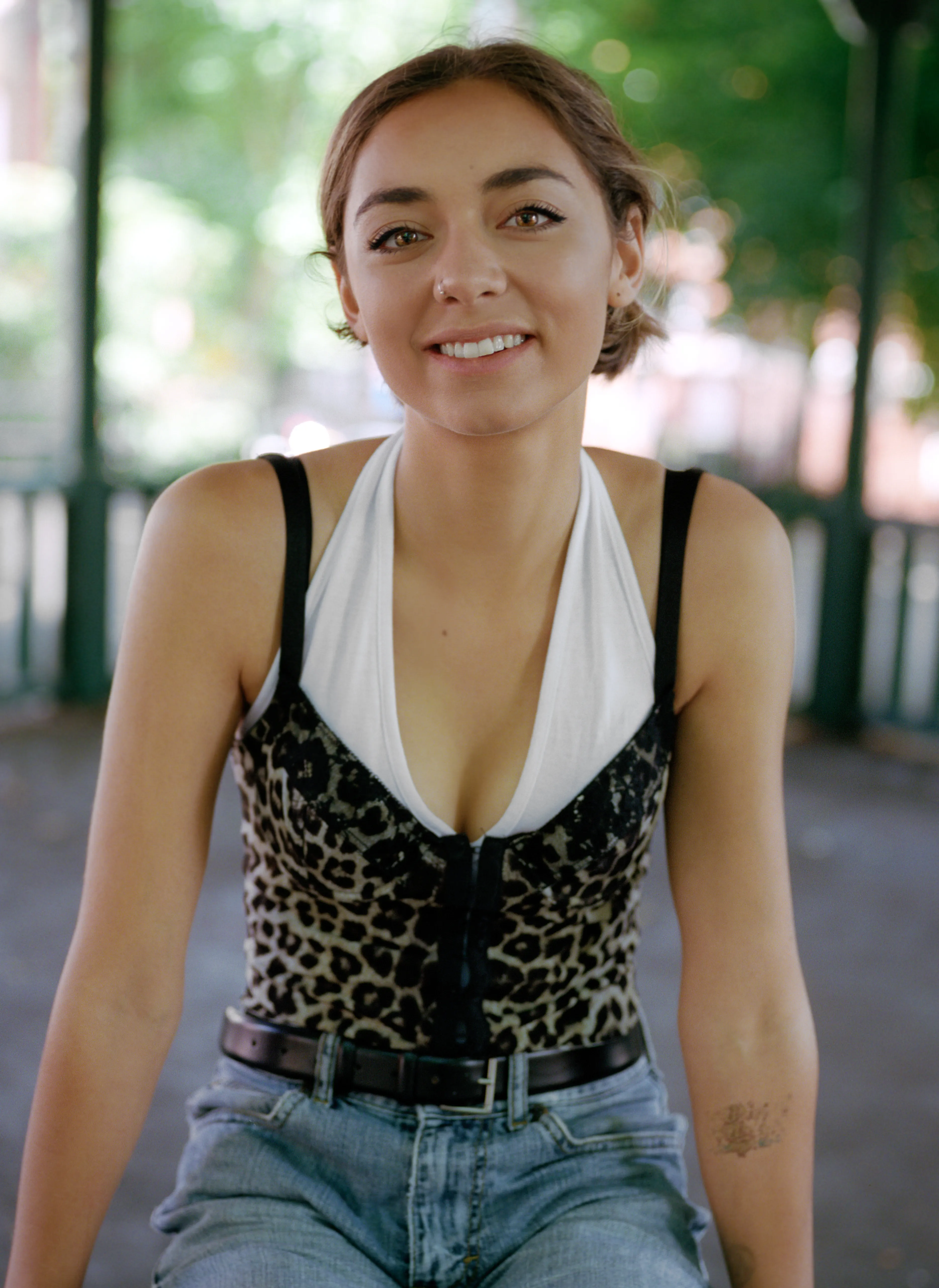 Riya laughs openly towards the camera, seated beneath a park pavilion with white railings and greenery behind.