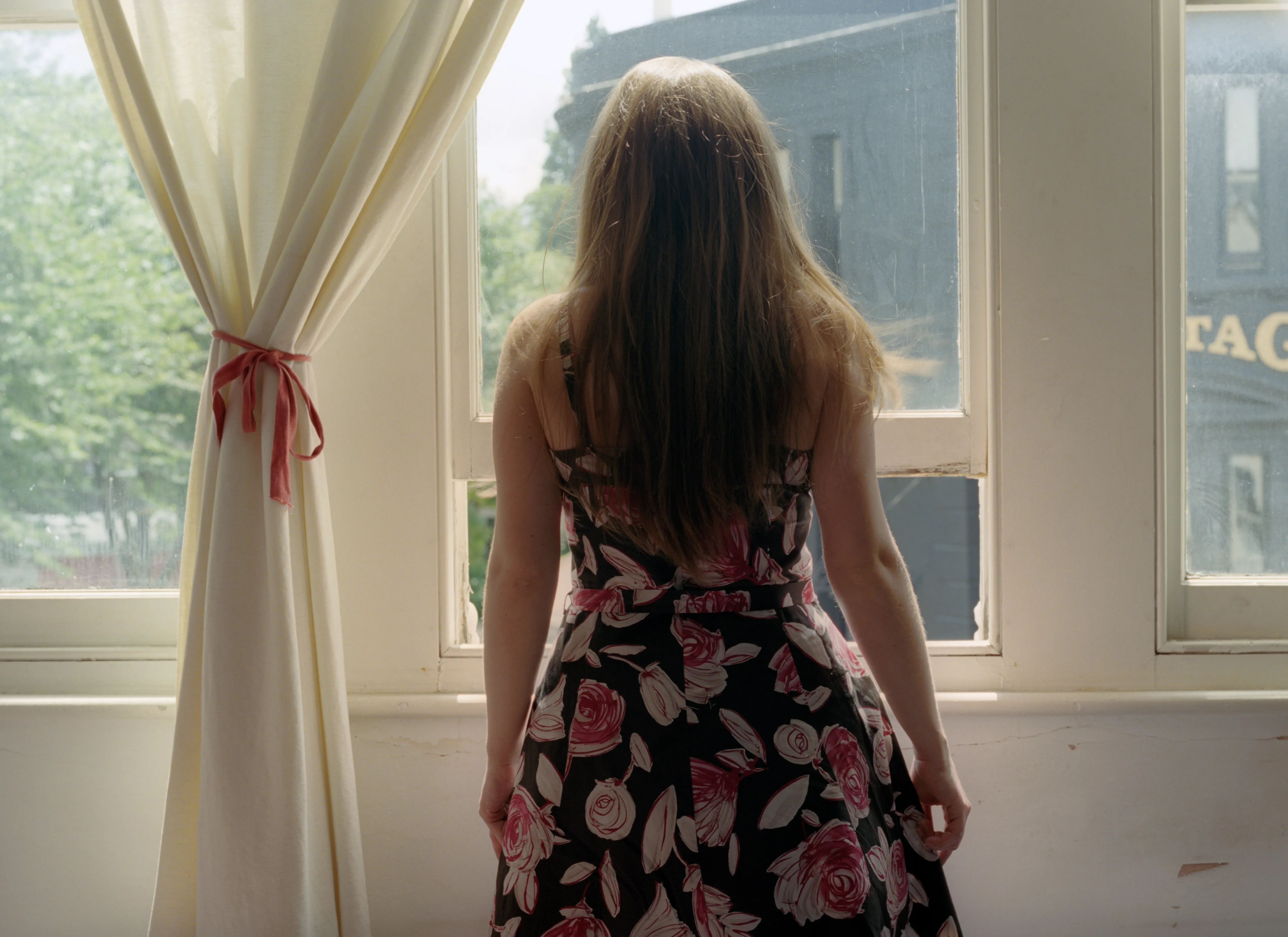 A woman in a floral dress stands with her back to the camera at a bright window, curtains tied back with ribbon, a street visible outside.