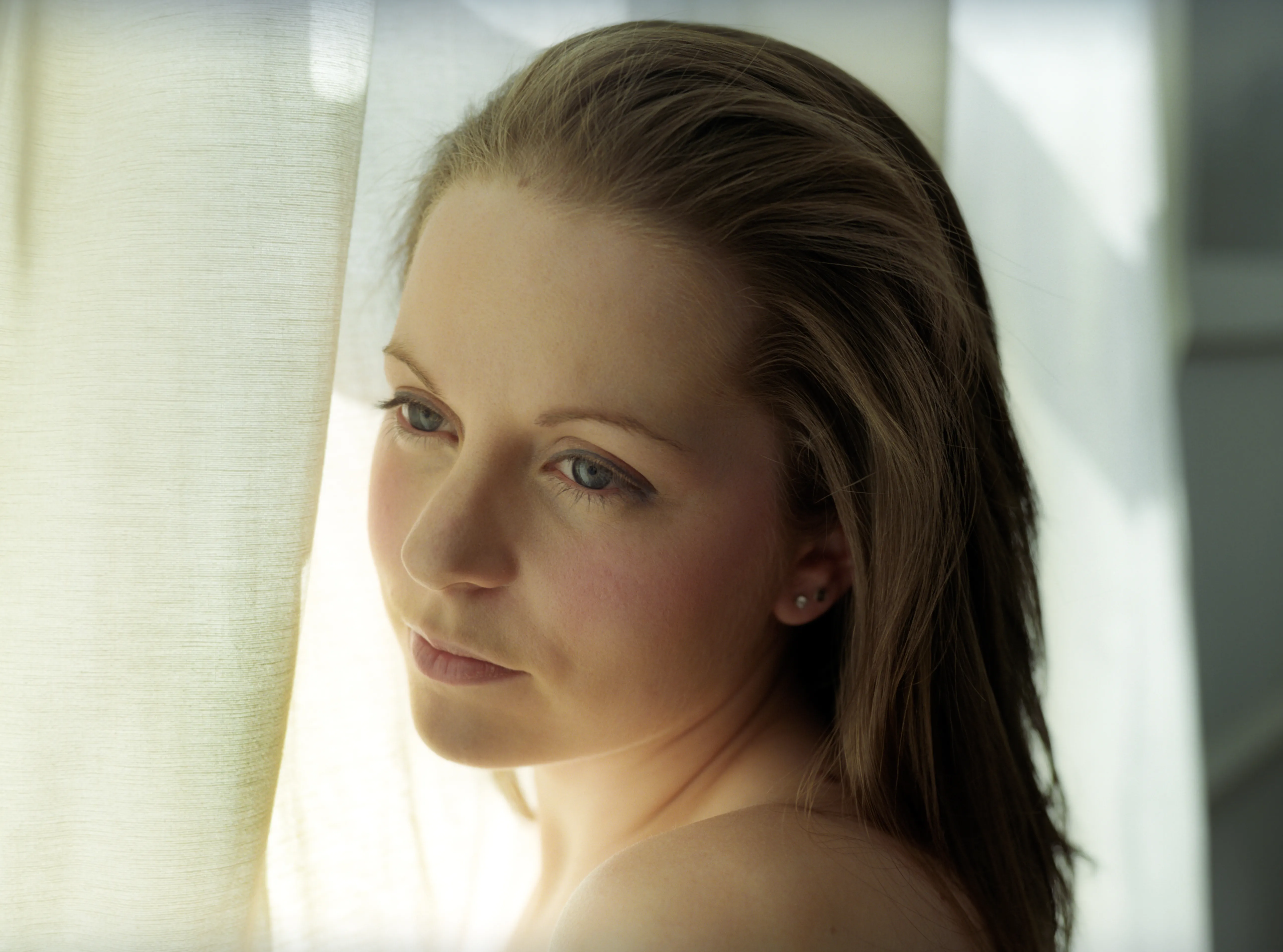 A bare-shouldered woman with her hair swept back looks toward the camera, lit by a sunlit sheer curtain behind her.