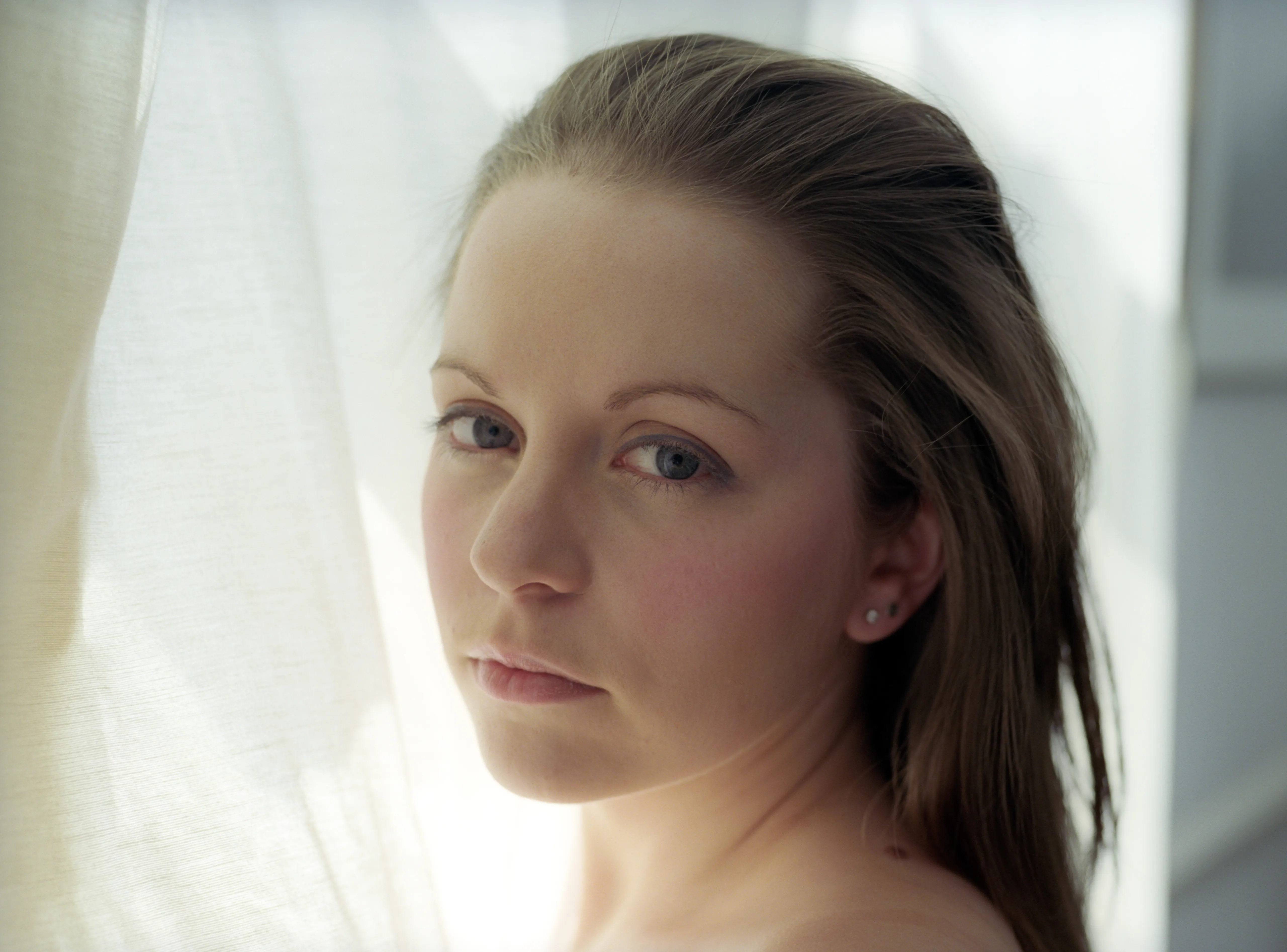 A bare-shouldered woman looks over her shoulder into the camera against a bright curtained window, her hair swept back.