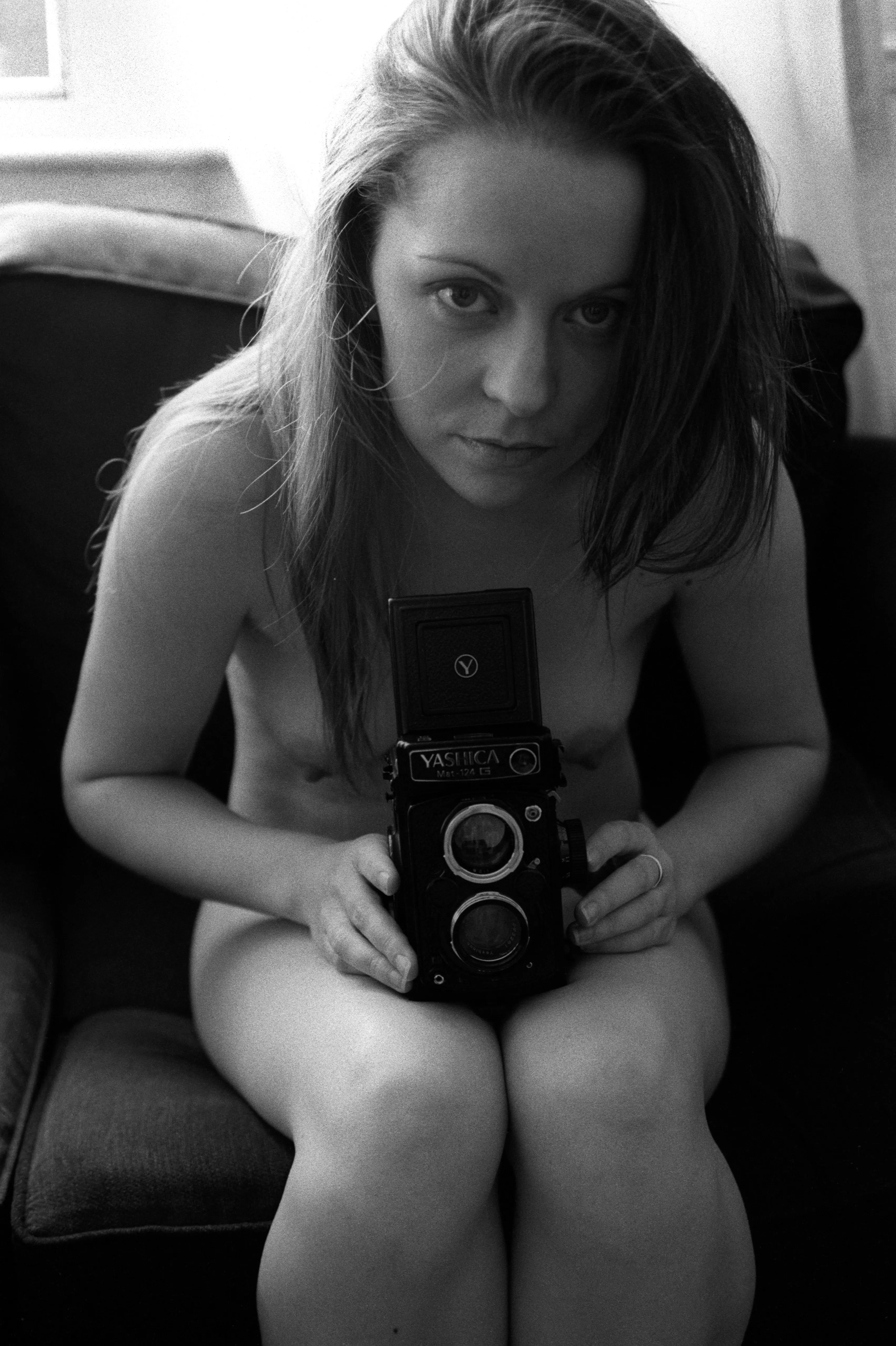 A seated nude woman holds a twin-lens reflex camera at her chest, looking over it toward the lens.