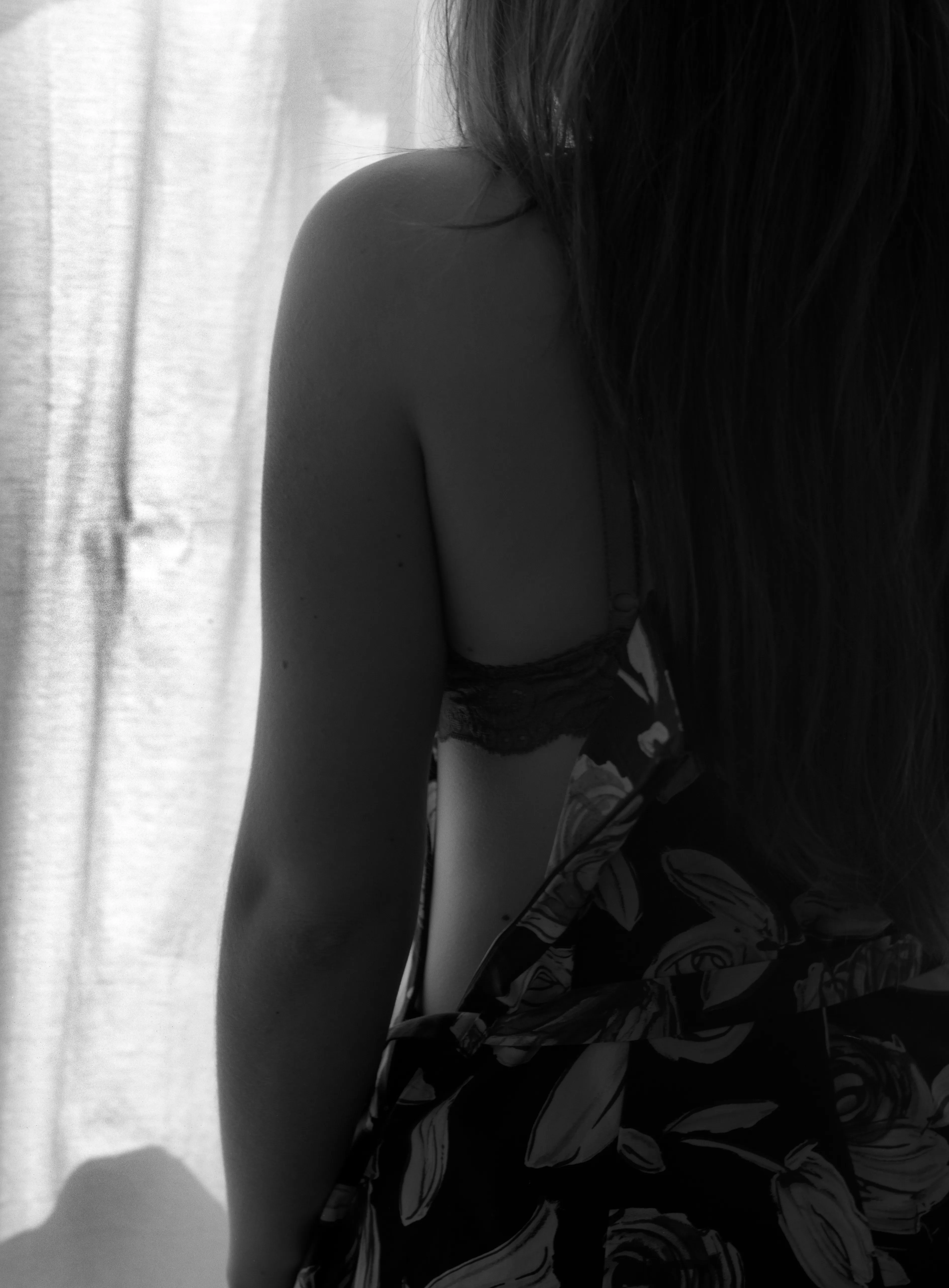 A close study from behind of a woman's bare shoulder and arm, a floral dress slipping from the shoulder to reveal a lace strap, against a bright curtain.