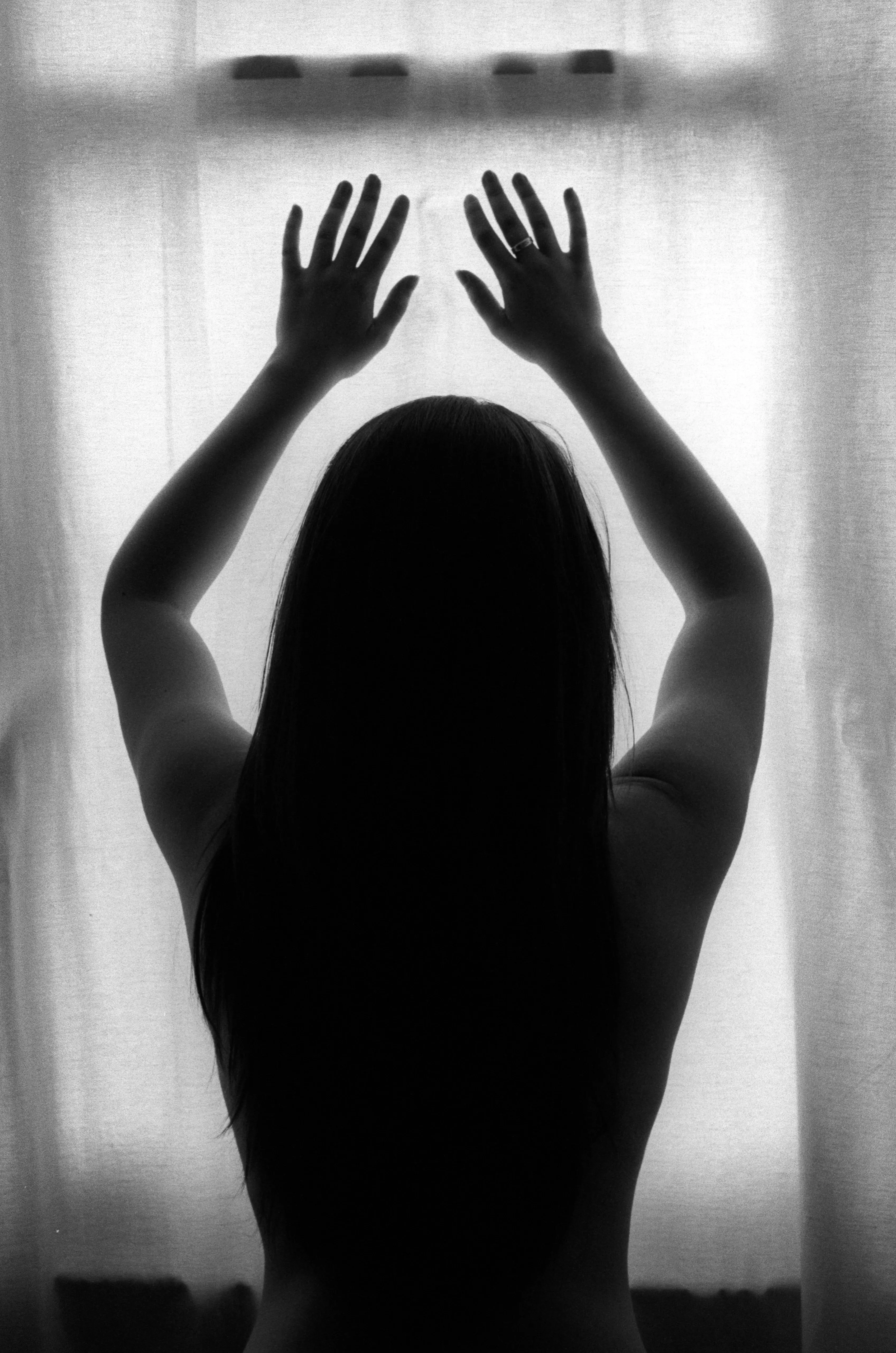 A nude figure in full silhouette presses both hands to a brightly backlit curtain, the body reduced to shadow.