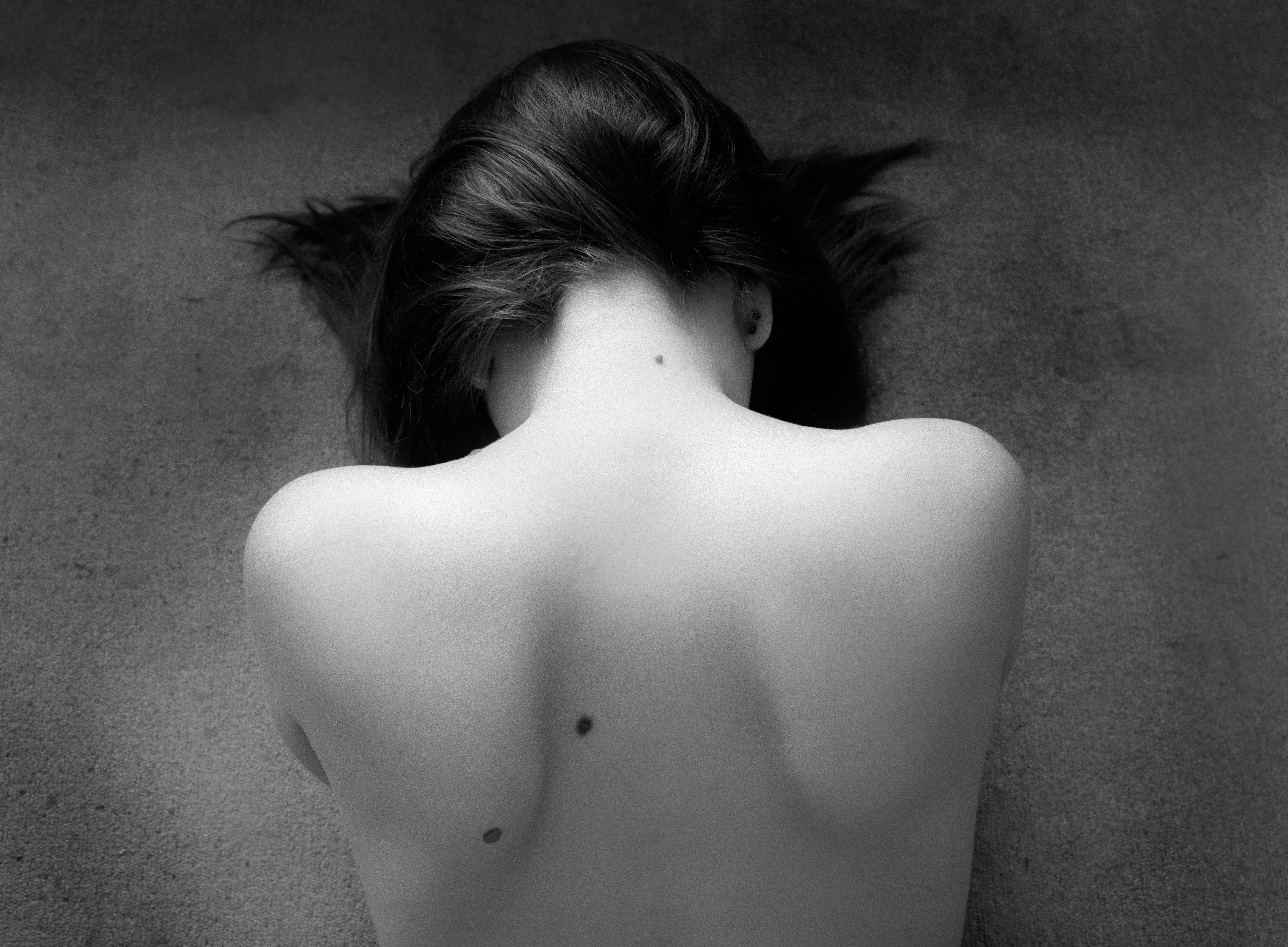 A woman's bare back seen from above, head bowed away, a single strand of hair tracing a line down her spine against a grey ground.