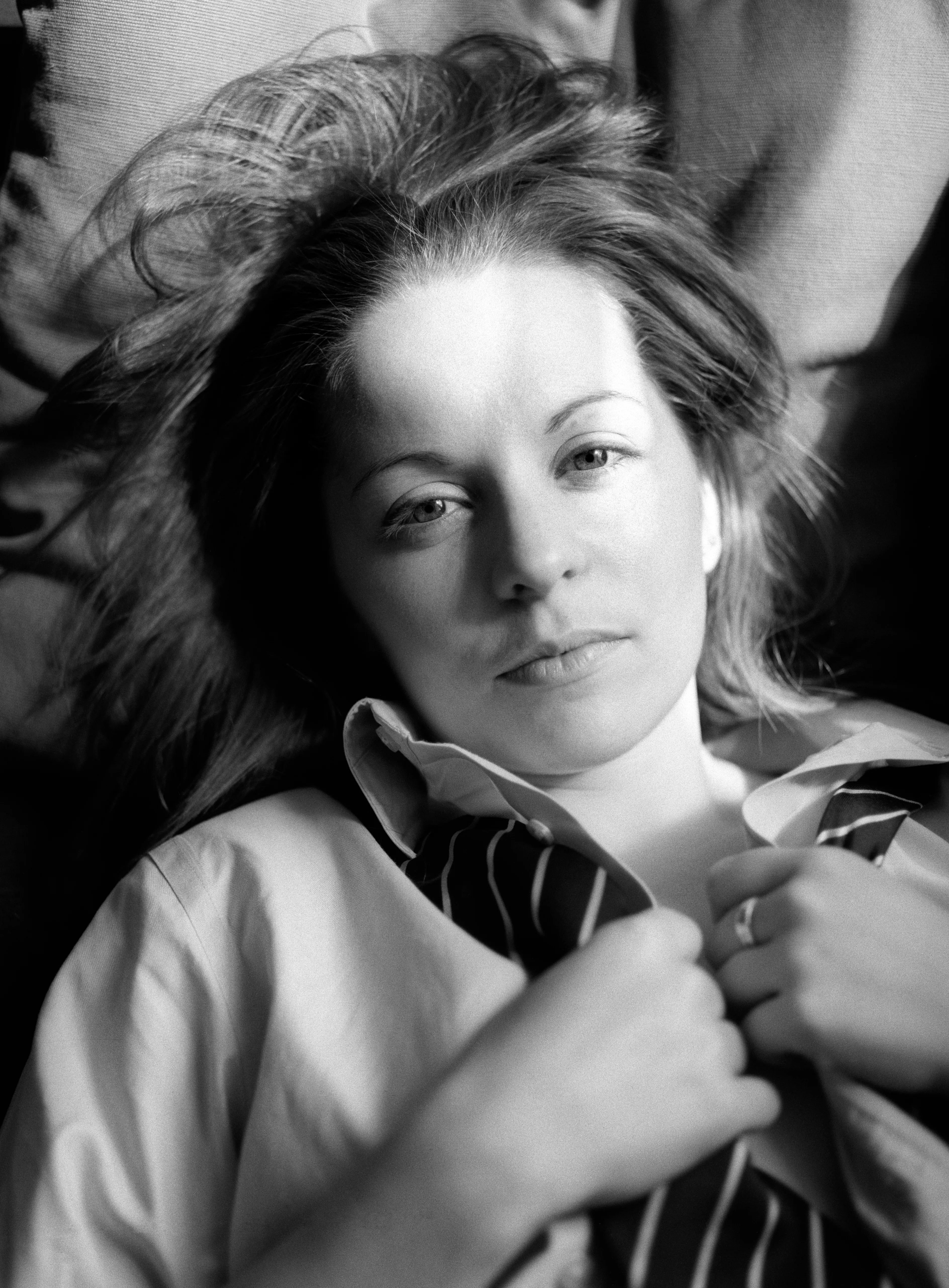 A woman lies with her hair fanned out, both hands drawing the open collar of a white shirt closed at her throat, looking up at the camera.