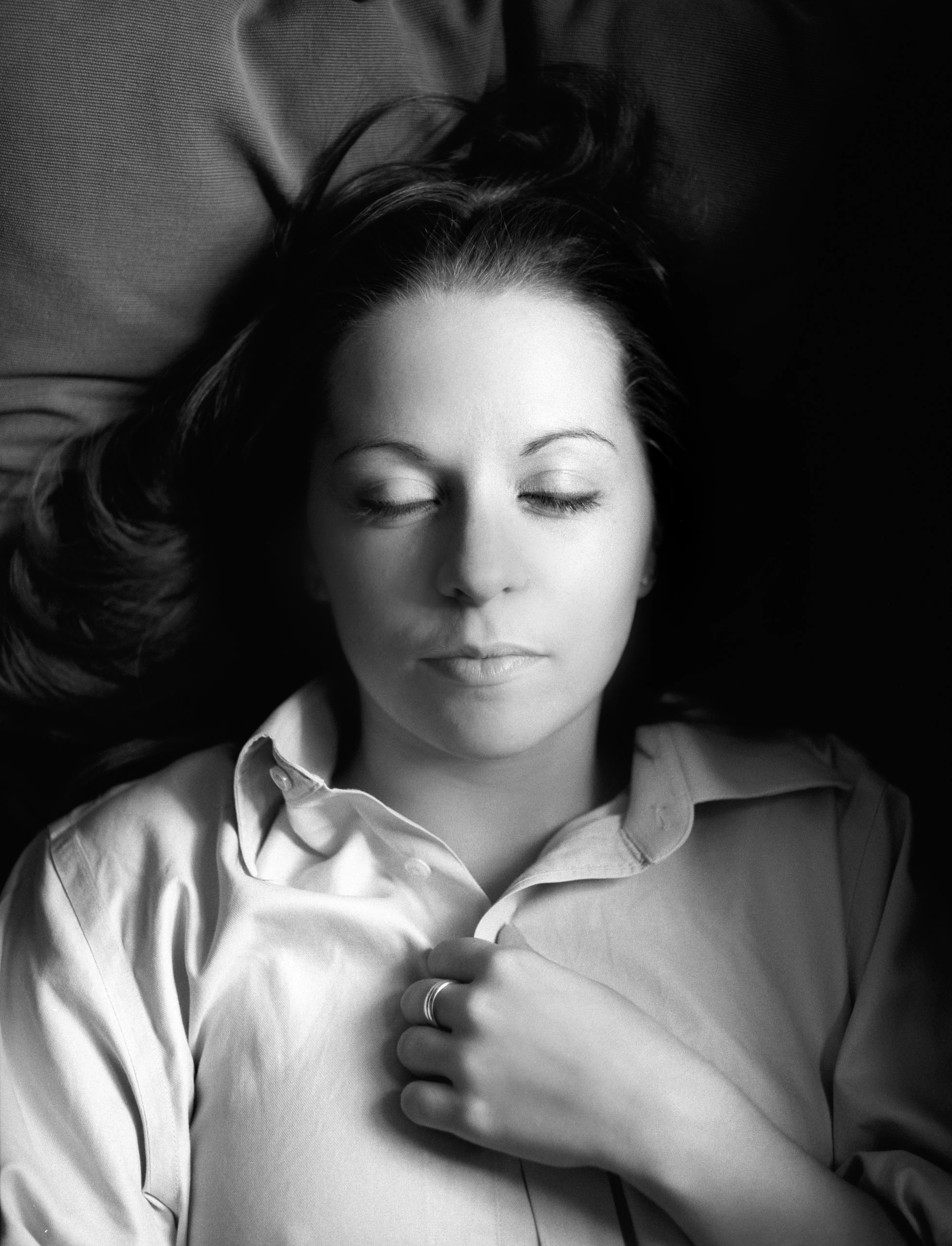 A woman reclines with her eyes closed in an open white shirt, her head turned on the pillow, hair loose.