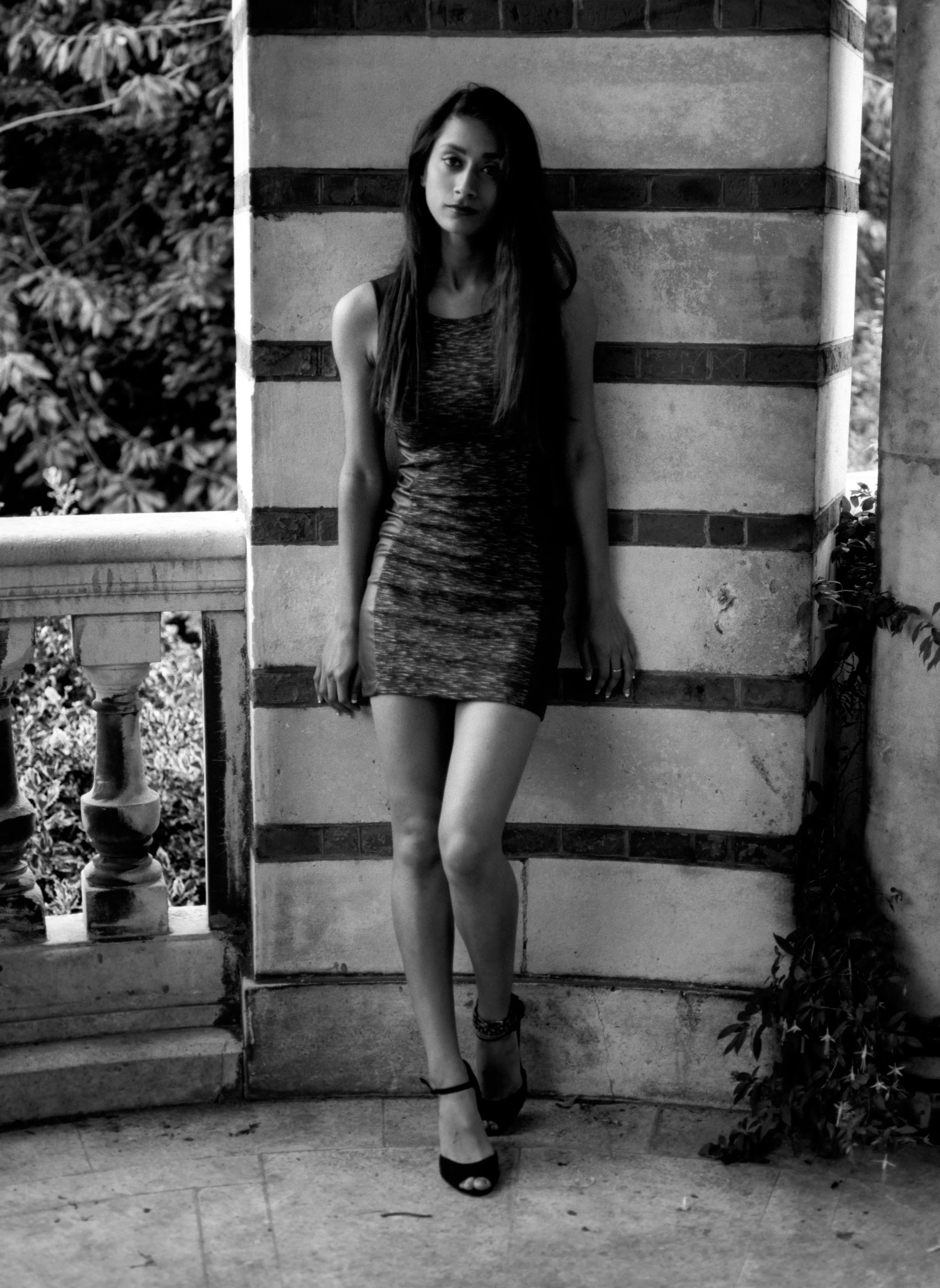 Shreena in a patterned dress, leaning against a banded stone pillar beside a balustrade, looking to one side. Black and white.