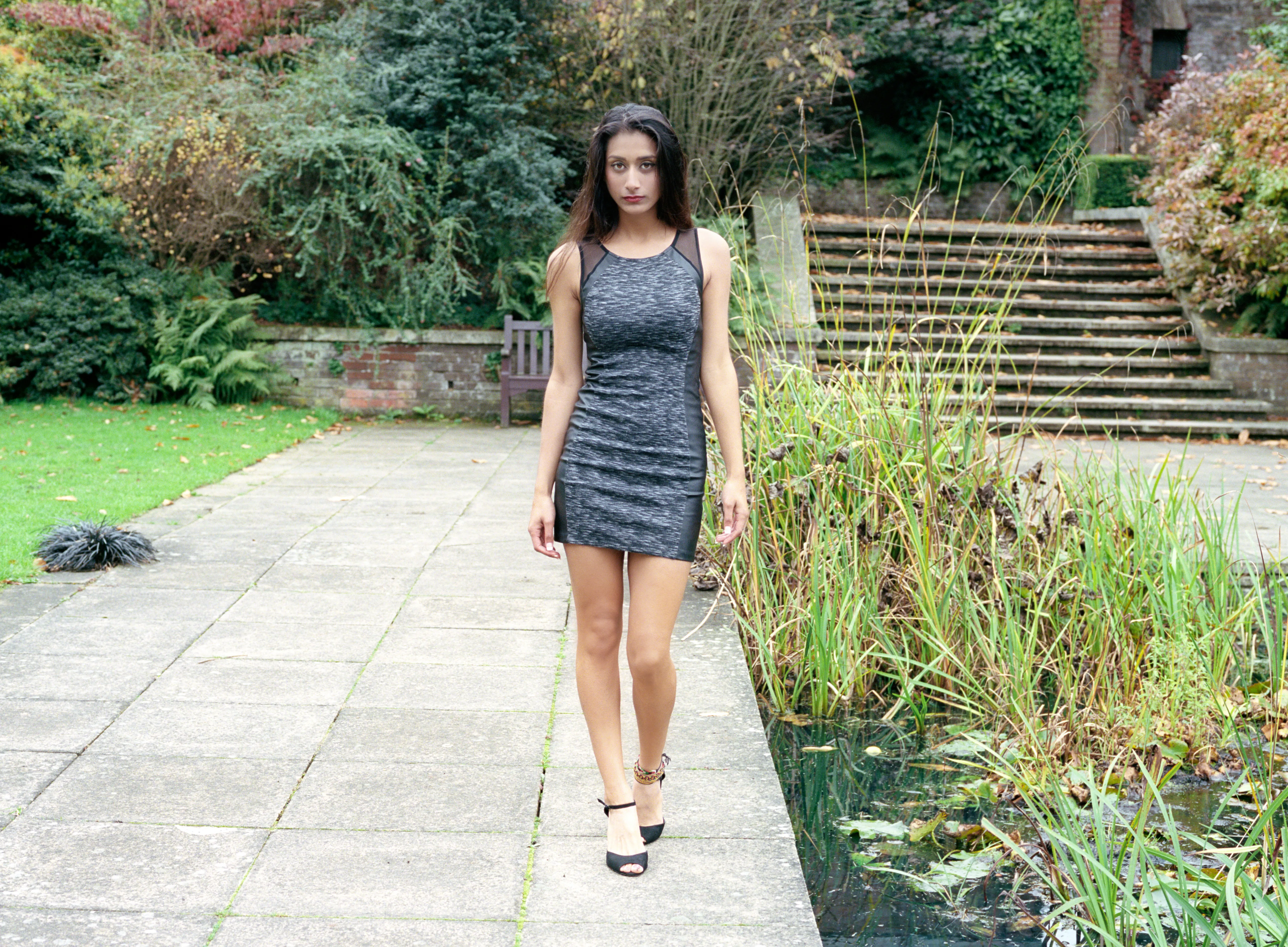 Shreena in a grey dress walking toward the camera along a stone garden path, steps and a pond behind her.