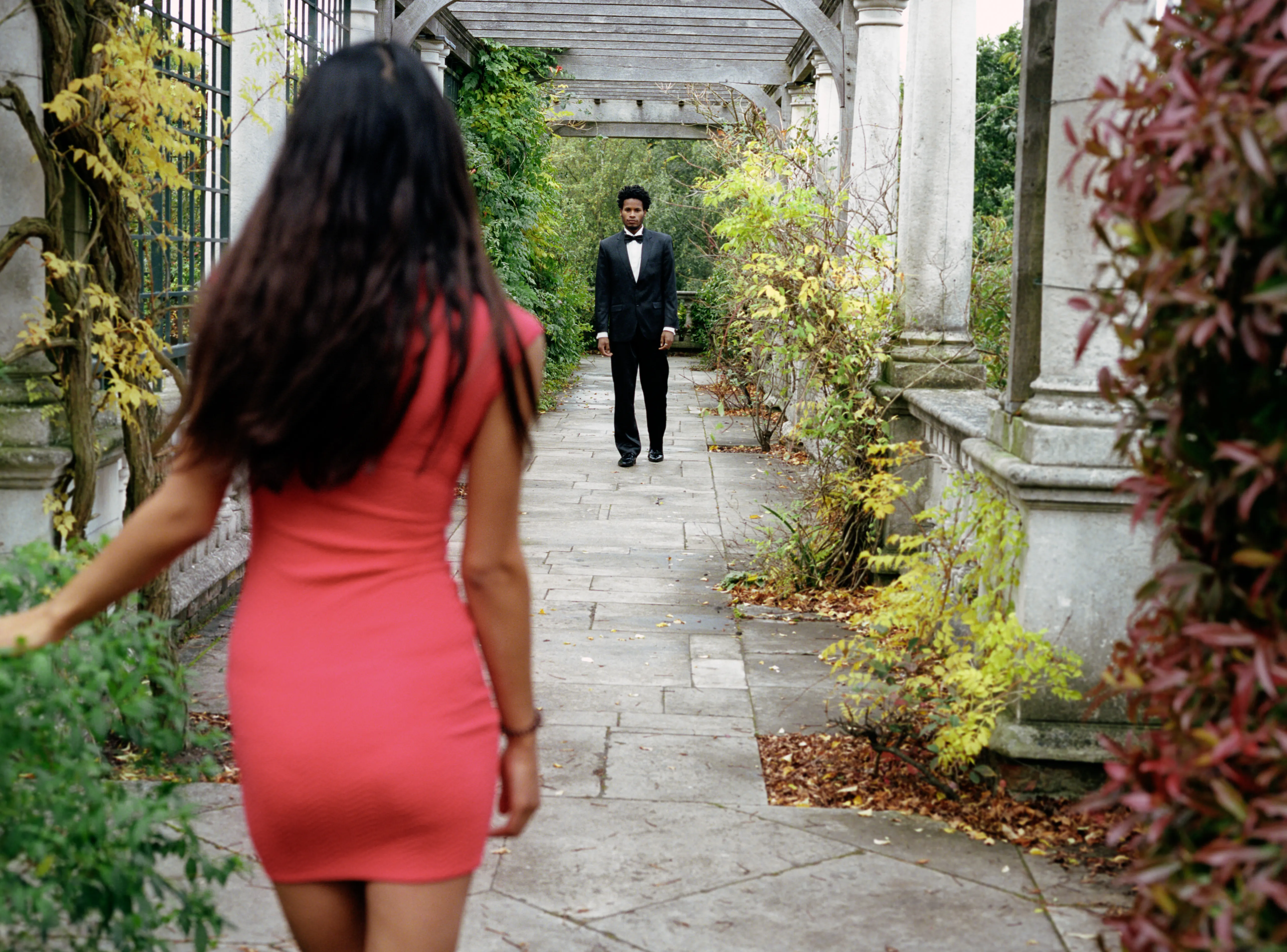 Shreena in a coral dress, seen from behind, facing Daniel in a dark suit who waits further down the pergola path.