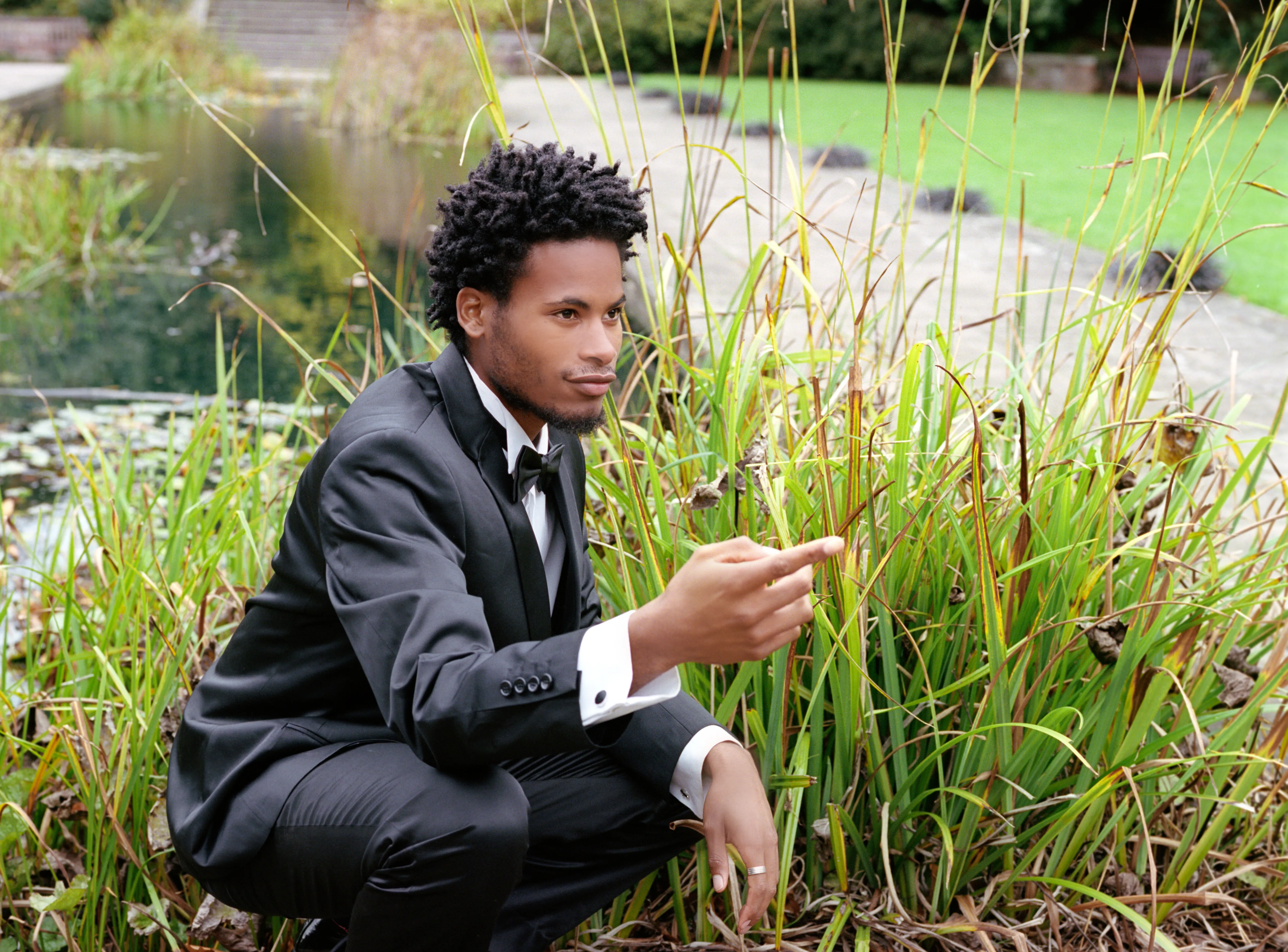 Daniel in a dark suit crouching among tall reeds at the edge of a pond, looking back over his shoulder.