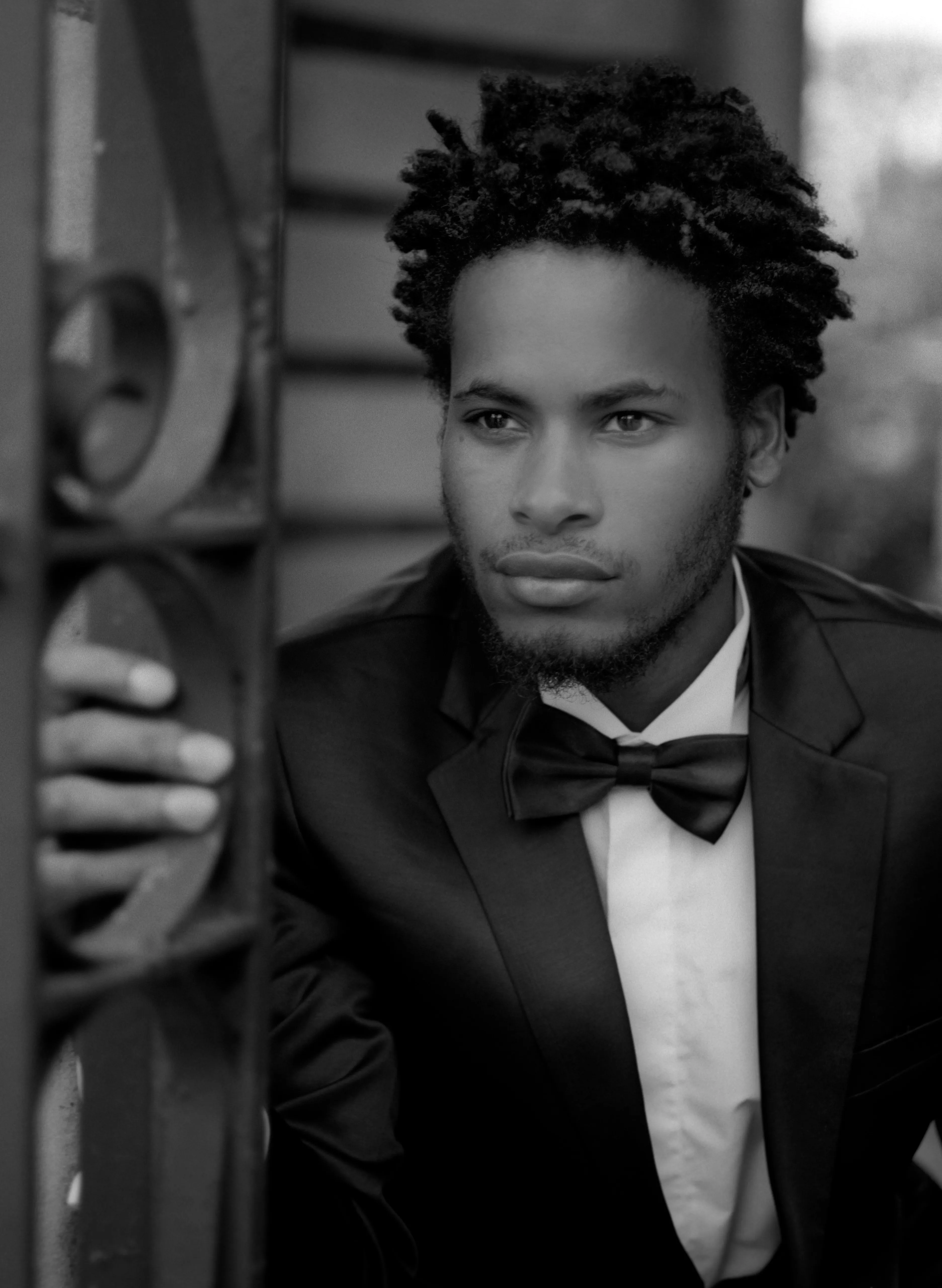 Daniel in a dinner suit and bow tie, head and shoulders, looking at the camera, one hand resting on an ornate iron gate, a slatted shutter behind him. Black and white.