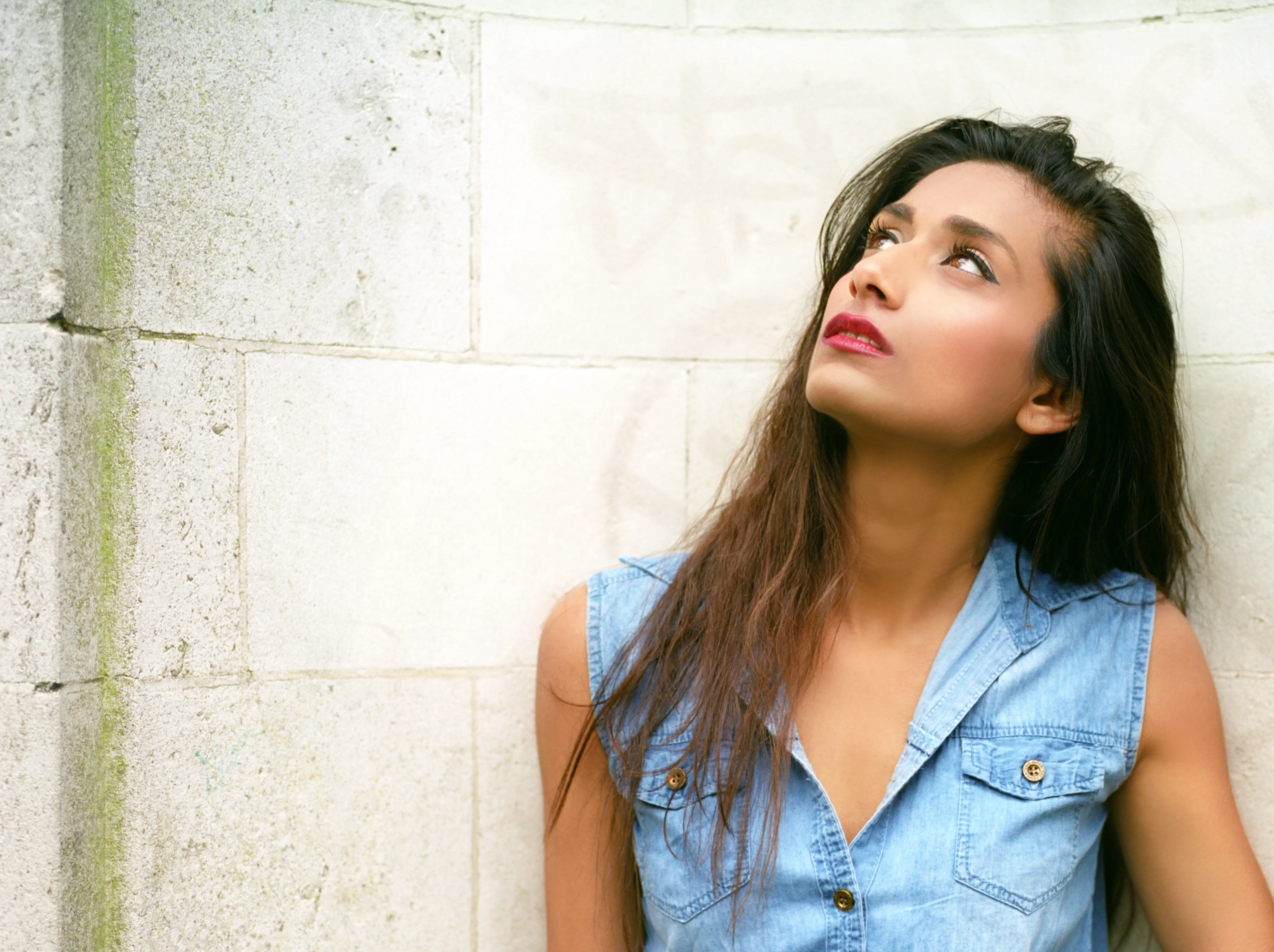 Shreena in a denim shirt against a pale stone wall, head tilted back, looking upward.