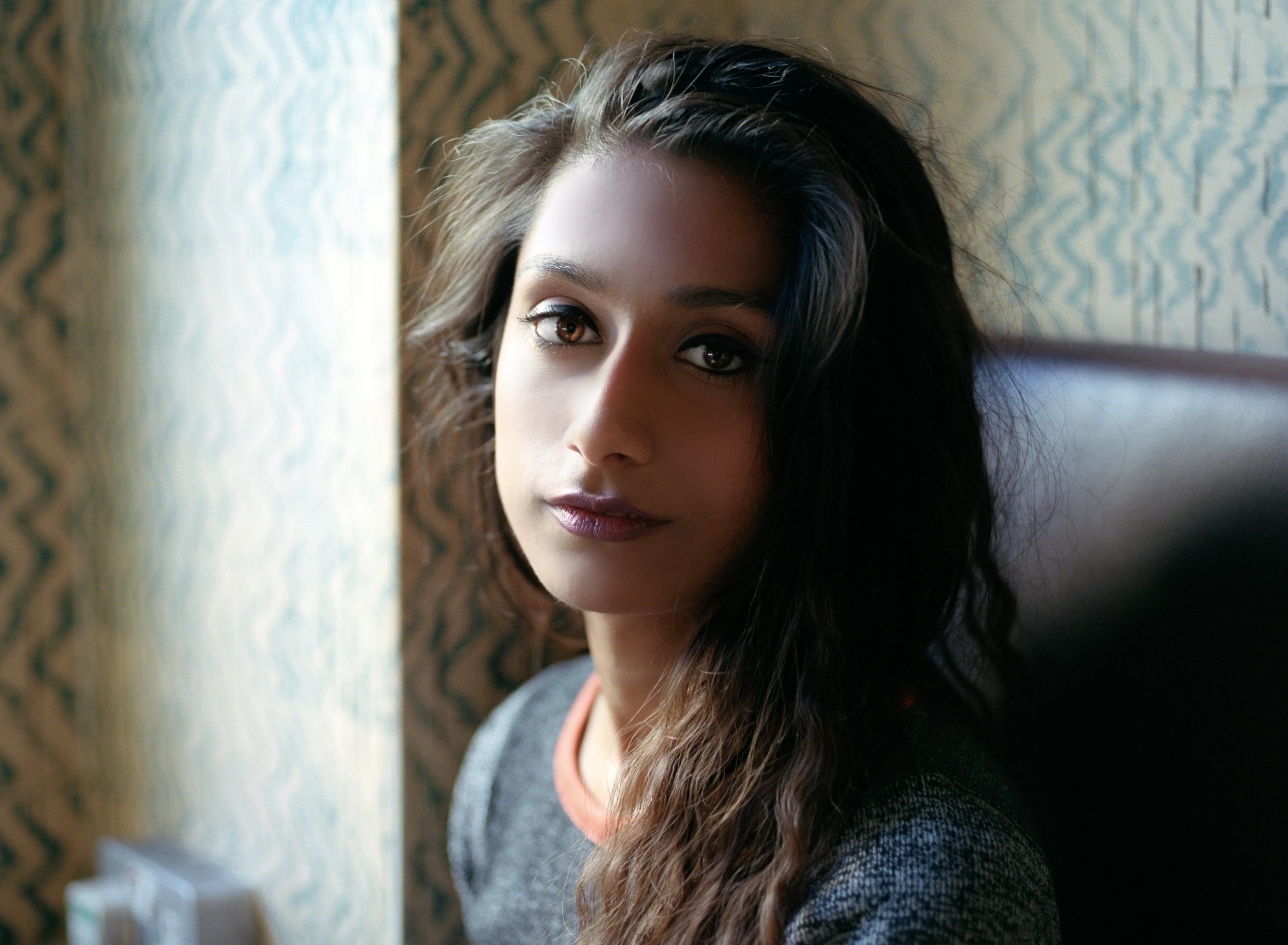 Shreena beside a window, hair loose, turned toward the camera in cool, dim light.