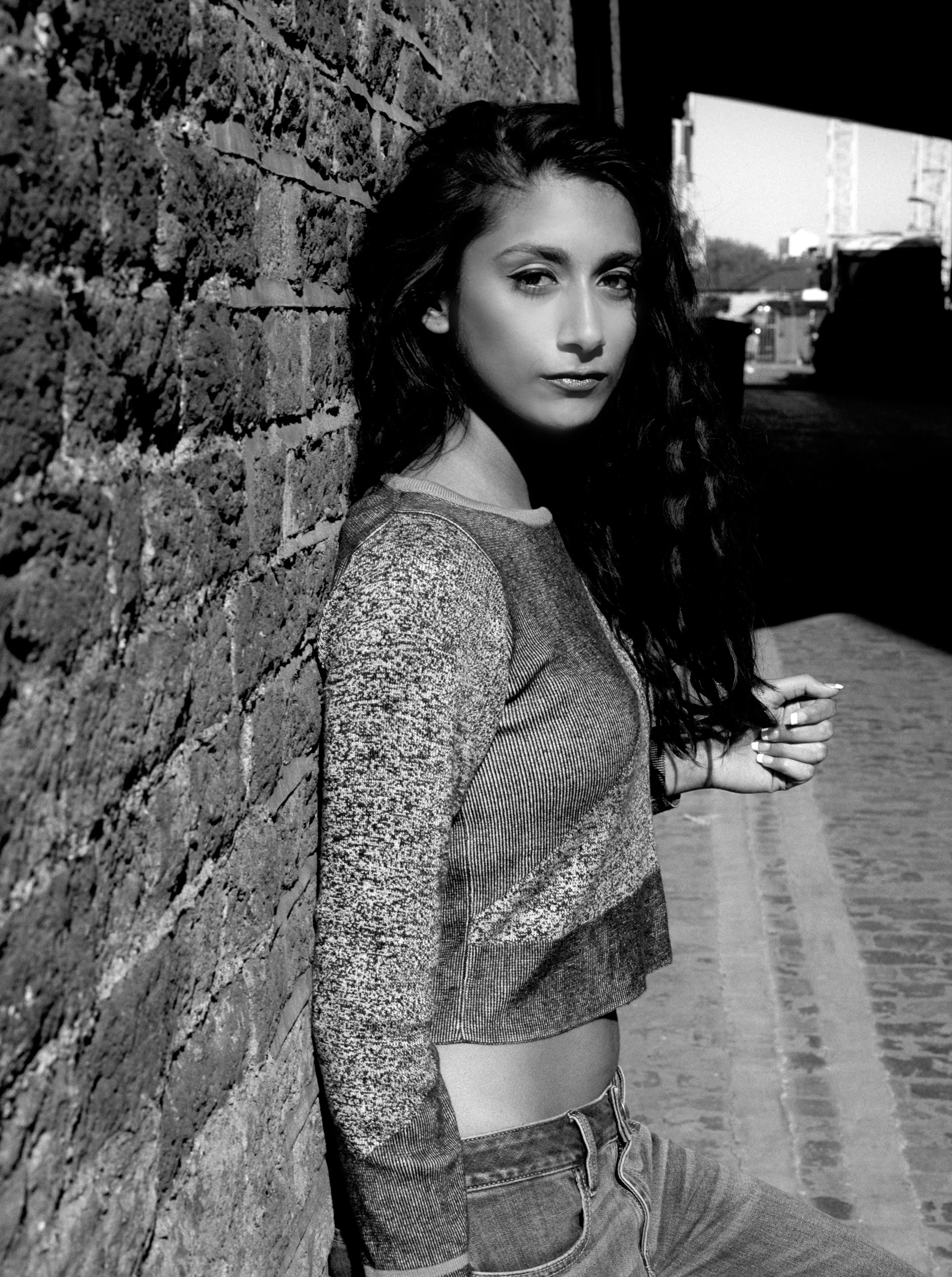 Shreena leaning against a rough brick wall, looking away down an alley. Black and white.