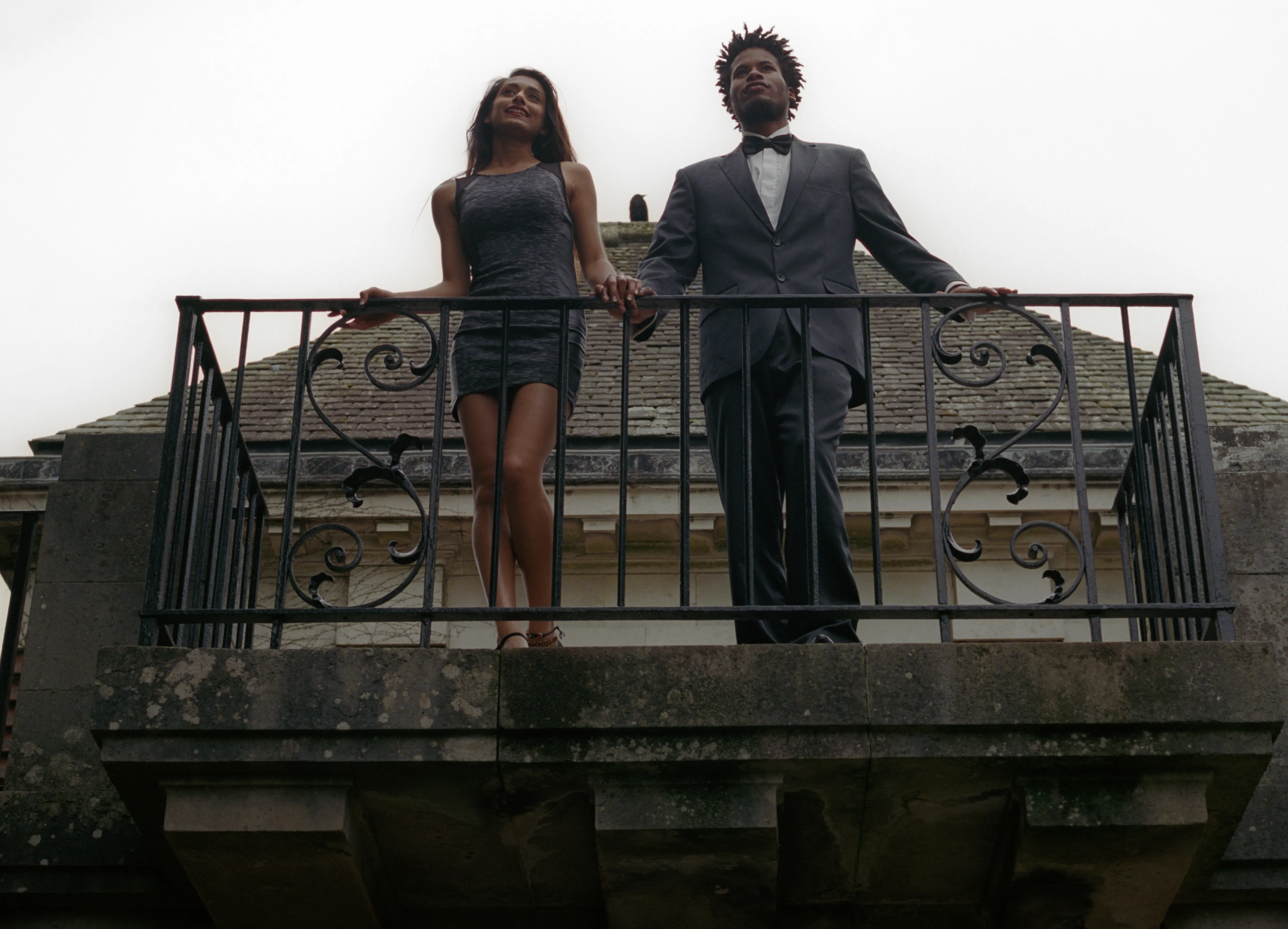 Shreena and Daniel on a stone balcony behind an ornate iron railing, seen from below against a pale sky, each looking in a different direction.