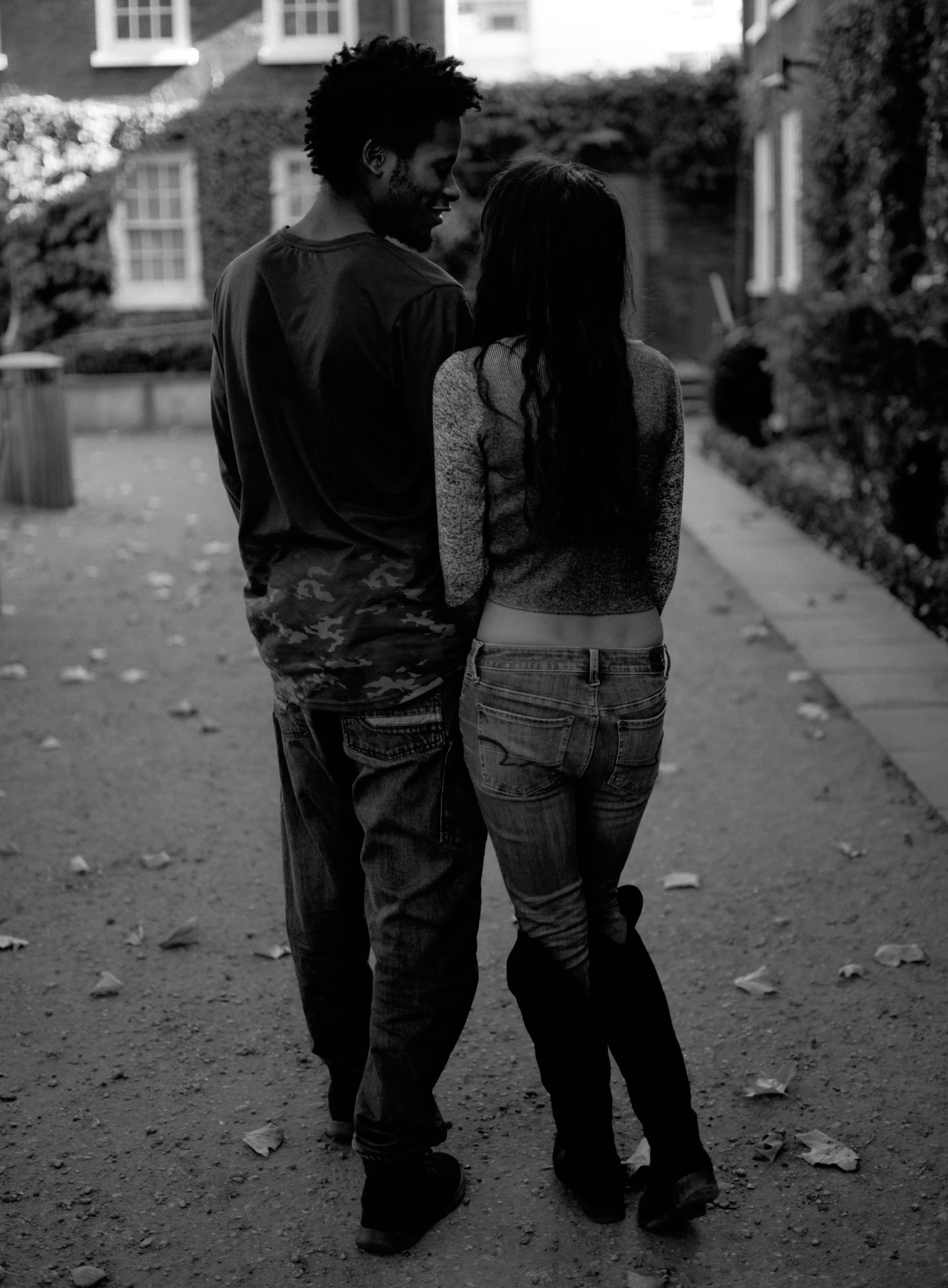 Shreena and Daniel walking away from the camera together along a path, arms around each other, an ivy-covered house behind. Black and white.