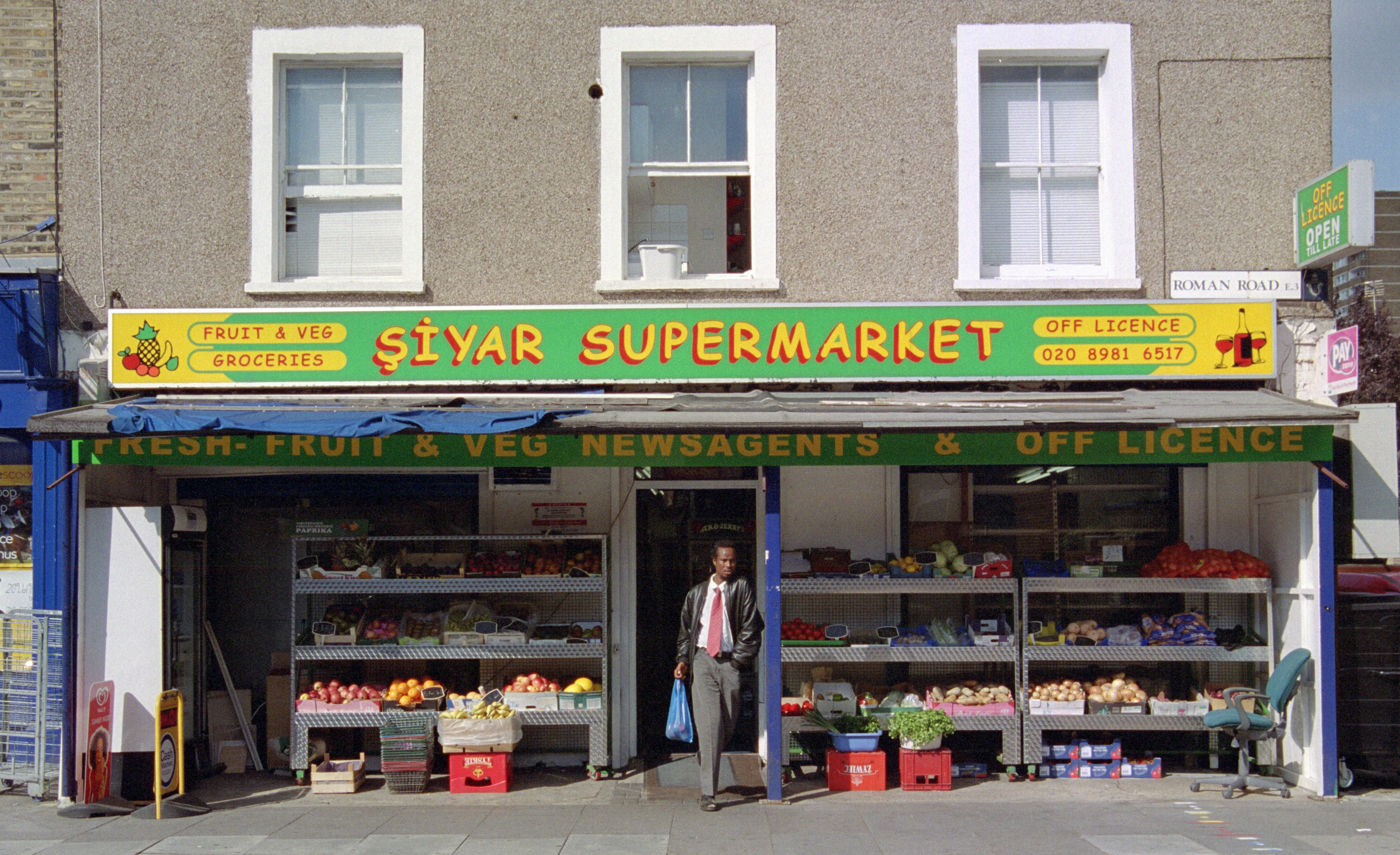 A man with a carrier bag in the doorway of Şiyar Supermarket, fruit-and-veg stalls flanking the green shopfront.