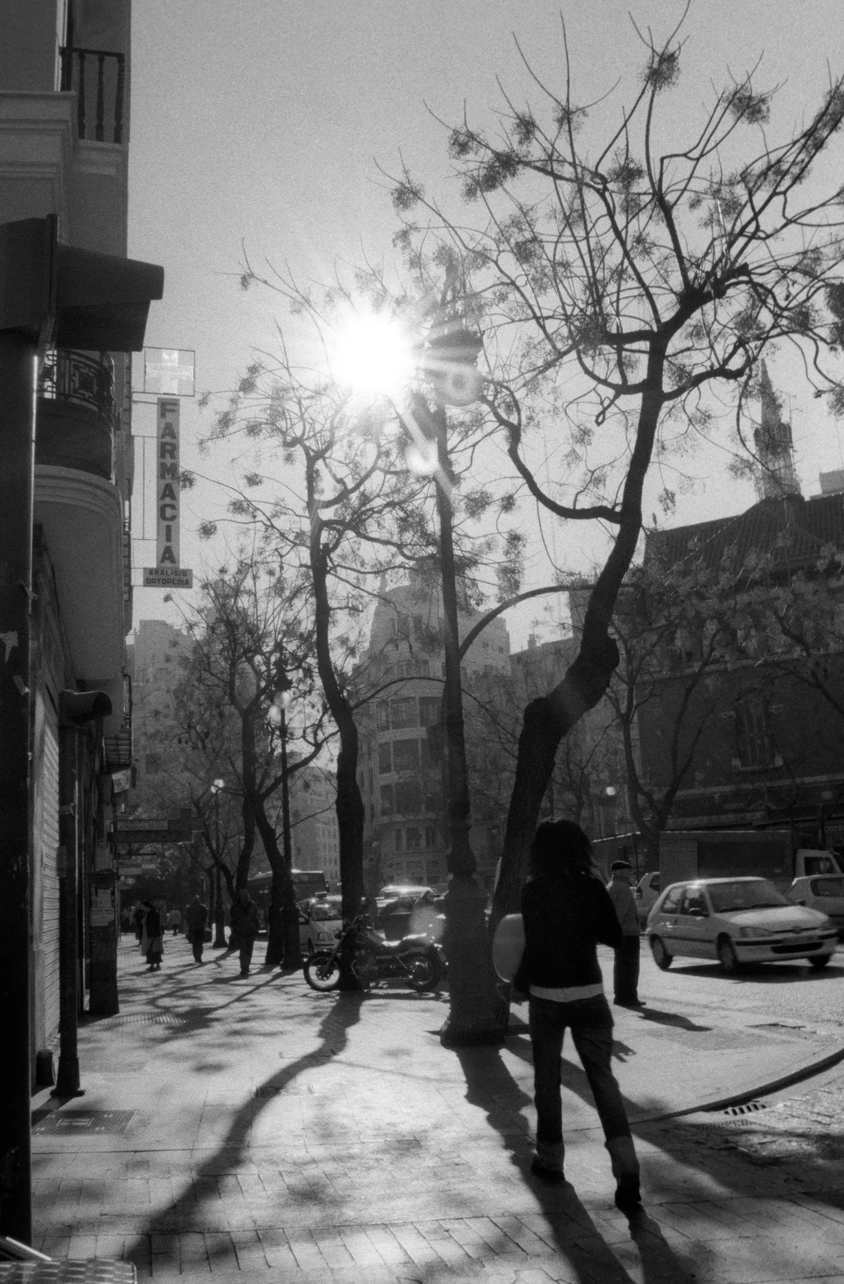 A woman walking into the low sun beneath bare trees, a pharmacy cross overhead, shadows streaming back.