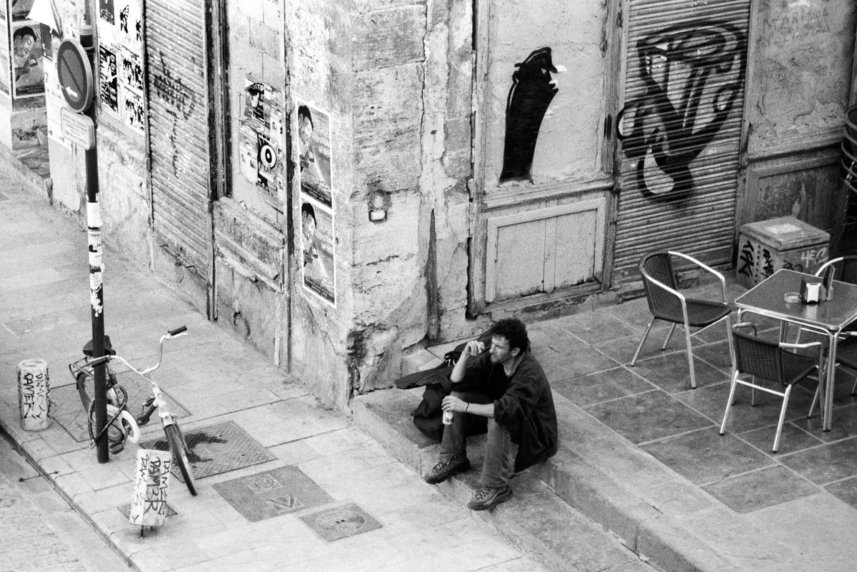From above — a man sitting on a corner step with a drink, a child's bicycle chained at the lamppost, café chairs to the right.