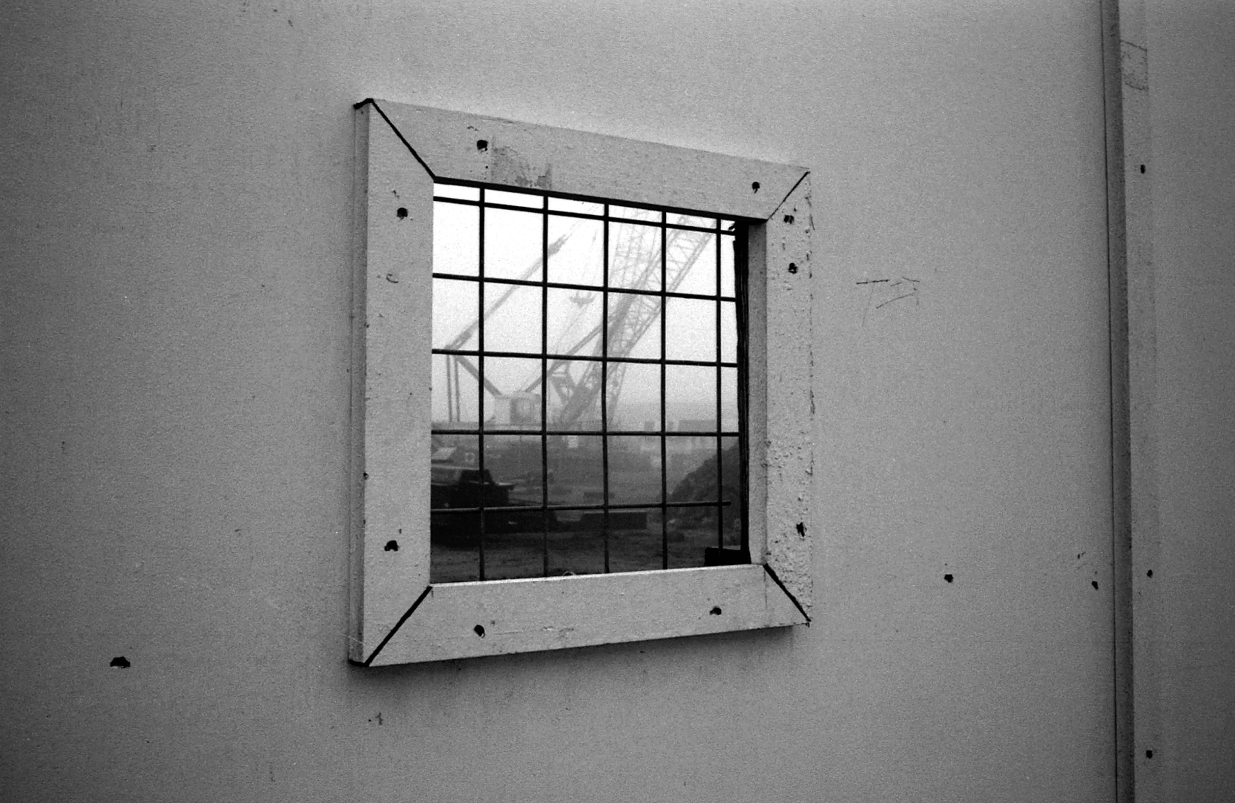 A crane and barges on the foggy Thames seen through a barred square window in a pale hoarding.