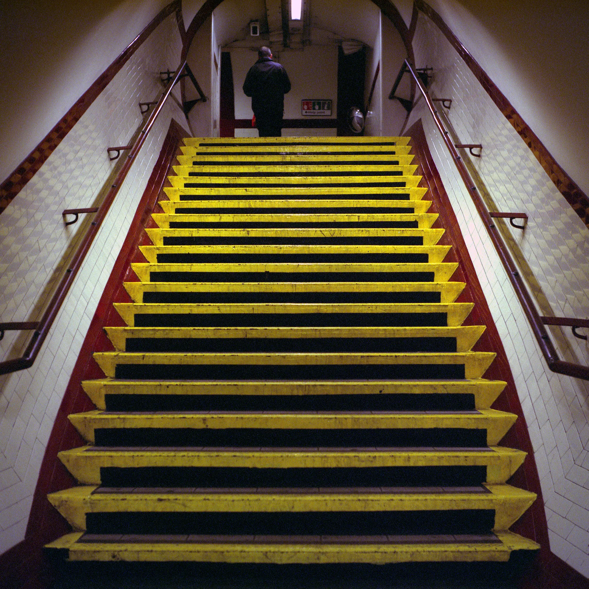 A figure crossing a London Underground platform behind the painted warning line; one of the two colour frames.