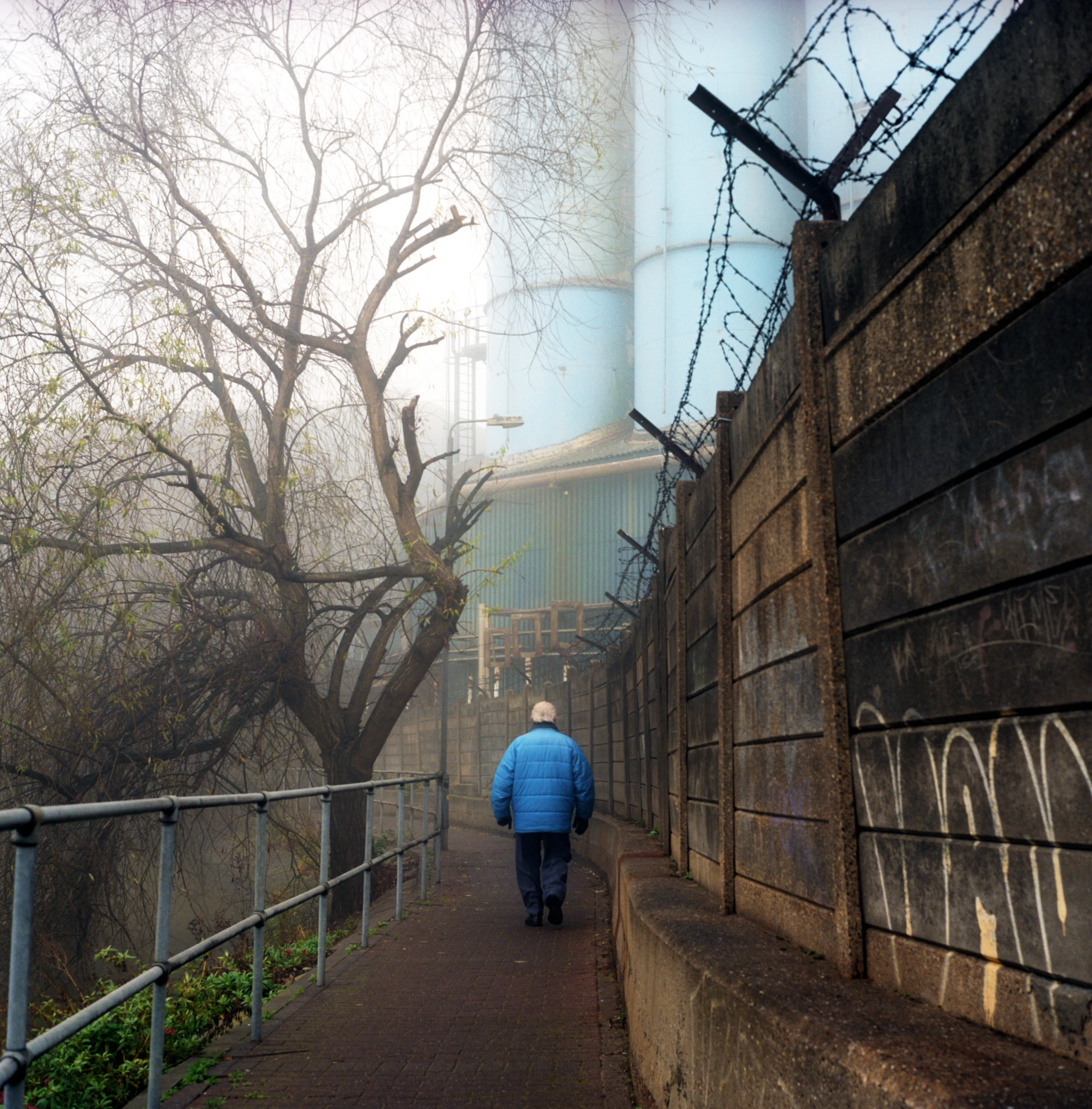 A solitary figure in a blue coat walking away down a misty riverside path, a bare tree and a barbed-wire-topped wall alongside.
